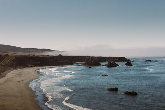 A serene coastal view with rugged stacks along a quiet shore under foggy, muted light.