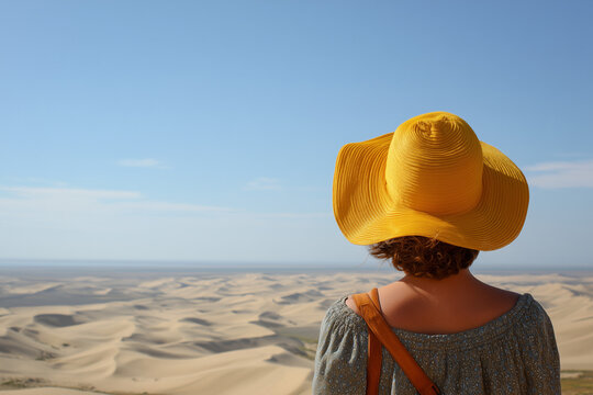 A woman in a yellow hat and shoulderless top overlooks a vast landscape of sand dunes. Calming blue sky