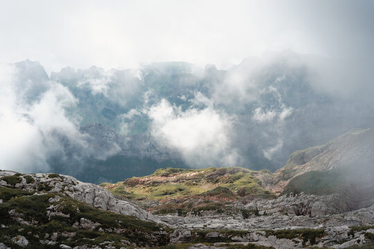 A sunlit patch of green and reddish ground contrasts with the rugged gray rocks as low clouds obscure the mountain cliffs in the distance.