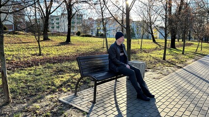 Man sitting on a park bench in winter clothing on a sunny day, surrounded by bare trees and long shadows