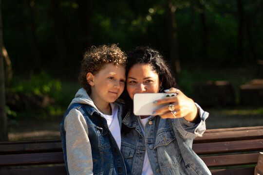 Young Caucasian mother and playful adorable little kid son making selfie photo or recording funny video, entertaining in public park