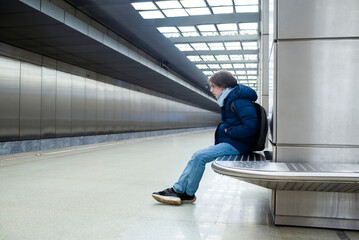 A teenage boy in a blue jacket sits on a bench on the platform, waiting for a train in the subway.
