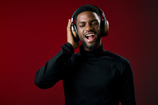 Young Black man in black turtleneck enjoying music with headphones on a vibrant red background, expressing joy and enthusiasm while listening to his favorite tunes