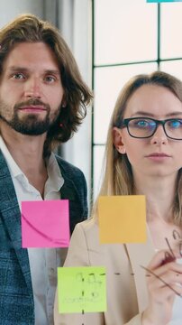Good-looking man and woman creators of design office brainstorming new ideas for joint project and recording marks on sticky notes posing on camera in modern office