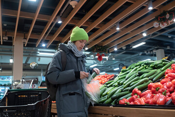 A teenage boy in winter clothes puts cucumbers in a plastic bag, getting ready to buy them. The child stands in the fruit and vegetable section of a modern grocery store.