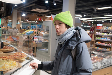 A teenage boy in winter clothes stands in front of a counter with pizza and other prepared foods, the child talks to the seller.