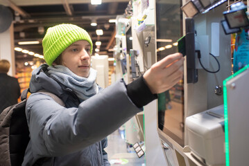 A teenage boy pays for purchases with a card at a self-checkout counter in a store. A child in winter clothes at a grocery store swipes a bank card at a payment terminal.