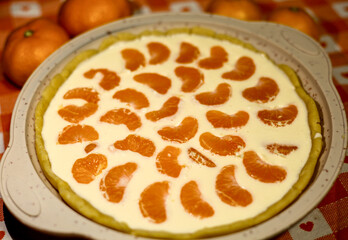 A round, homemade tart with a creamy custard filling and a decorative pattern of fresh mandarin orange segments, ready to be baked or served. Sweet dessert for holidays.