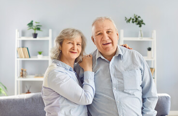 Portrait of a happy senior family couple wife and husband standing in the living room at home hugging and looking cheerful at camera. Love, care and relationships in retirement concept. .
