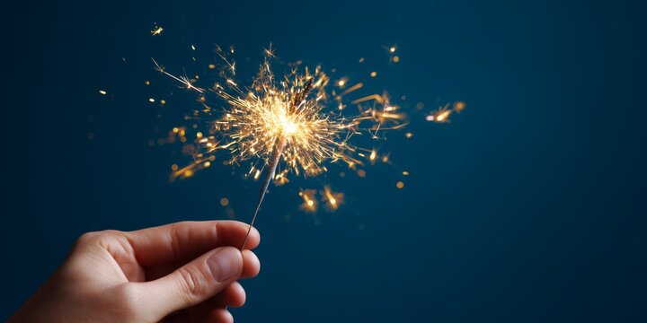 Candid close-up studio shot, a hand holding a lit sparkler with golden sparks flying, against a smooth seamless midnight blue background.