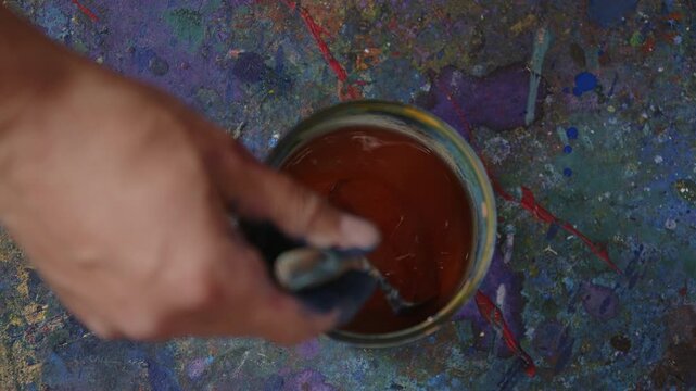 Hand r&uuml;hrt orangene Fl&uuml;ssigfarbe im Eimer um &ndash; k&uuml;nstlerischer Mischprozess mit Pigmenten, kreativer Studioumgebung und dynamischer Farbbewegung