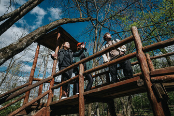 A group of friends stands on a rustic wooden bridge in a sunlit forest, looking out toward the sky. Outdoor adventure, nature, and camaraderie highlight active moments.