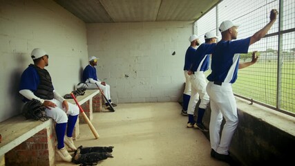 Baseball players in blue uniforms wait and watch intently from the team dugout, ready for their turn on the field. Their equipment is ready nearby.