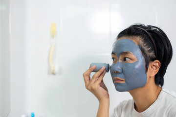 Asian woman applies clay facial mask in front of bathroom mirror. Skincare routine promotes clean skin and natural beauty. A refreshing beauty treatment for detoxifying and rejuvenating the face.