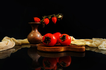 Autumn still life: ripe persimmons on a wooden board, a physalis branch in a vase, dark background