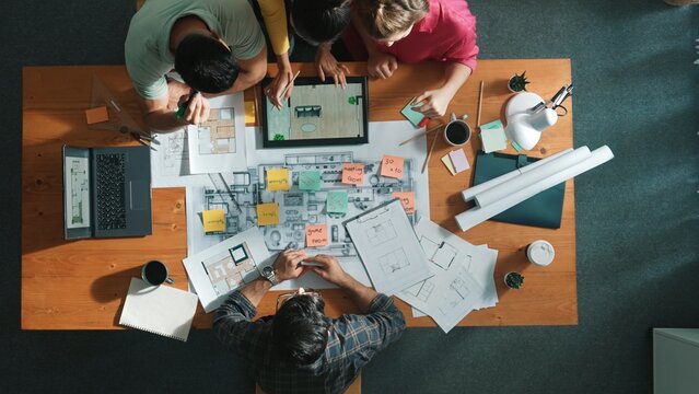 Top down view of manager writing at blueprint while marketing team plans design. Aerial view of architect engineer working together sharing idea and taking a note in project plan at table. Symposium.