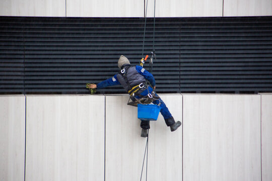 Industrial climber hangs from the roof of building, cleaning the building ventilation grille at height. This dangerous work process takes place in challenging weather conditions