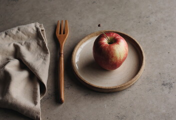 A single red apple rests on a plate with a wooden fork beside it, evoking a simple, rustic still life.