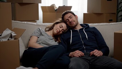 Tired young couple is peacefully napping on the couch amidst stacks of cardboard boxes. The joyful but fatigued new homeowners are pausing their unpacking efforts to recharge