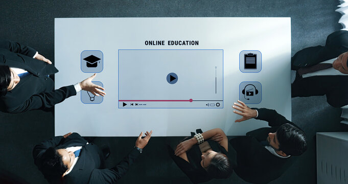 A diverse group of professionals engages in a collaborative discussion about online education, utilizing a digital interface with a video player and educational icons on the table. Trope