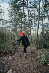A hiker wearing an orange cap and green jacket walks up a forest trail, using trekking poles. The scene conveys outdoor adventure, focus, and natural solitude amid tall pines and autumn leaves.