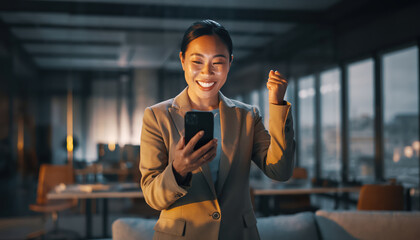Happy businesswoman celebrates success while using smartphone in modern office setting. warm lighting and contemporary design create inspiring atmosphere