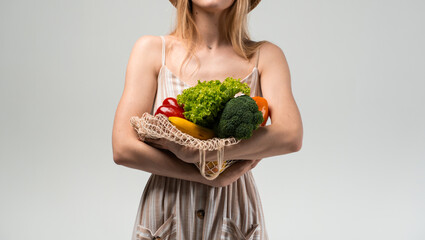 Woman in striped dress holding eco mesh bag with fresh vegetables in hands on light background