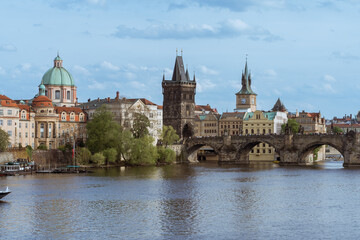 Historic view of Charles Bridge, Old Town Bridge Tower and riverside buildings in Prague on a clear spring day, with calm Vltava River and city architecture in soft natural light.