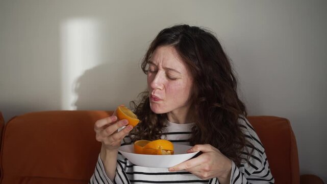 Pregnant woman experiencing morning sickness and nausea in her early pregnancy, discovering that eating a juicy orange helps alleviate her symptoms as she relaxes on the sofa in her cozy living room
