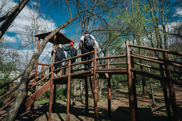 A group of friends and family walk along a raised wooden bridge through a lush forest park. Bright blue sky, green trees, and backpacks convey an active outdoor adventure.