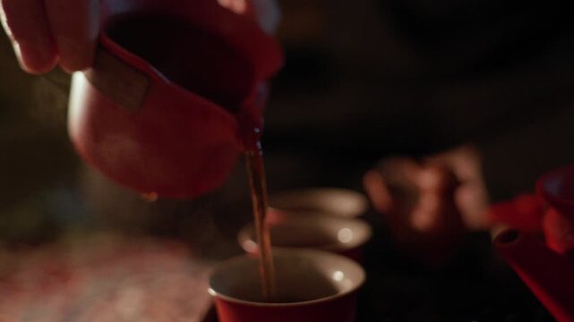Tea is poured from a red teapot into cups and over a figurine during a tea ceremony, with drops, splashes, bubbles, and steam visible