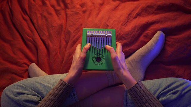 Young woman sitting cross-legged on red blanket, playing green kalimba with gentle movements, creating serene musical atmosphere while exploring meditative sound and personal artistic expression