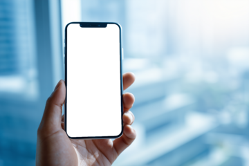 A close-up view of a hand holding a smartphone with a blank black screen inside a bright modern office environment. The blurred background highlights the phone, making it ideal for technology concepts