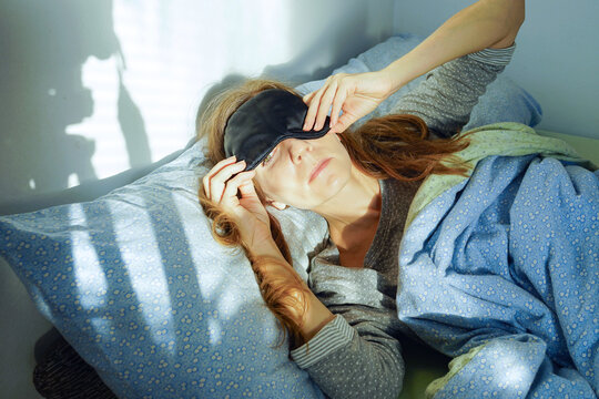 A woman lying in bed pulls a sleep mask over her eyes to block out sunlight during a migraine attack. Representing hormonal imbalance, cyclical sensitivity, endocrine stress,  and chronic fatigue.