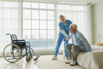 Rehabilitation healthcare. Physiotherapist nurse helps old man patient in wheelchair stand up walking. Man in chair for people with disability doing few steps with nurse helping. Medical caregiver