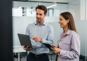 Two smiling colleagues collaborating on a laptop and tablet in a modern office environment discussing a project