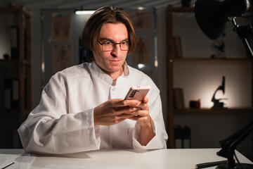 Shocked Surprised Doctor in Glasses Holding Smart Phone in the Clinic on Night Shift sitting at Desk