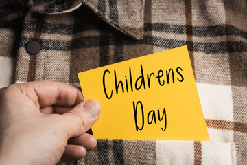 Memory of Children's Day: A hand holding a bright, orange and yellow note with the inscription "Childrens Day" on the background of a men's shirt with a brown and beige check