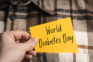 World Diabetes Day: A hand holding a yellow note with the inscription "World Diabetes Day" against the background of a casual brown and white plaid shirt, promoting health awareness.