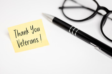 Appreciation: Yellow note "Thank You Veterans!" Symbolizing gratitude, lying next to an elegant pen and glasses on a clean, white desk top.