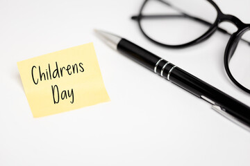 Important Dates in the Calendar: A yellow sticky note with the inscription "Childrens Day" lying on the white desk top, next to black-rimmed glasses and an elegant pen.