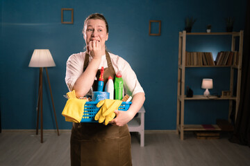 Portrait of unhappy woman in apron holding bucket with cleaning supplies biting nails, nervous and...