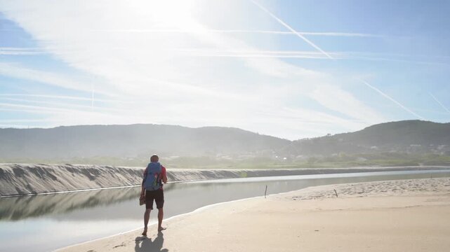 Lonely backpacker man walking along sandy beach washed by Atlantic Ocean waves in Portugal in low sunrise light. Shot during the Camino Portuguese &mdash;  popular pilgrimage route. Slow motion footage.