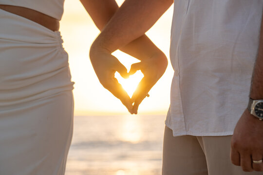 Close up silhouette of couple hands making a heart shape against a golden sunset beach background. - Powered by Adobe