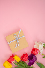 Gift box with white ribbon next to multicolored tulips on a pink background