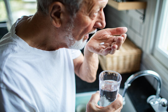 Senior man focused taking medication with water at home kitchen