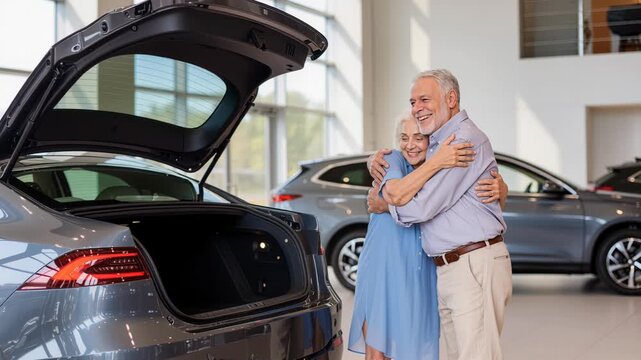 Two elderly Caucasian seniors share joyful moment in modern car dealership, arms wrapped around each other as they smile, with new vehicles trunk open, displaying its interior., family happiness