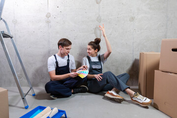 House decoration DIY renovation concept. Happy couple sitting on floor in empty room of new home planning design. Husband and wife holding swatch palette choosing colors for painting wall in new house