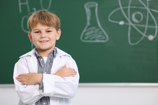 Smiling little boy in laboratory coat near green chalkboard with formulas indoors, space for text. Child and science - Powered by Adobe
