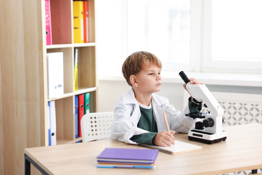 Cute little boy with microscope writing at desk indoors. Child and science